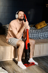 Wall Mural - Sleeping young man listening to music in headphones and cleaning floor of bedroom with vacuum cleaner while sitting by his bed