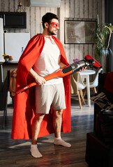 Wall Mural - Young man in red mantle and mask of superhero holding electric vacuum cleaner while standing in the center of living room at home