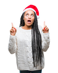 Poster - Young braided hair african american girl wearing christmas hat over isolated background amazed and surprised looking up and pointing with fingers and raised arms.