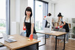 © sofiko14 - Portrait of Caucasian young woman holding sponge, detergent cleaning on table in office room looking at camera. Multicultural team in aprons and red gloves cleaning shelves, windows, floor in office.
