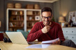 © Jelena - A smiling adult male college teacher reading and grading exams from his students while sitting in front of a laptop.