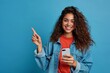 Young happy excited latin woman pointing at mobile phone isolated on blue background. Smiling female model holding cellphone using cell presenting advertising new trendy application, Generative AI