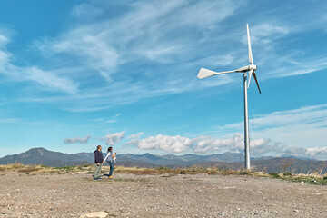 Wall Mural - Green energy concept. Couple with dog staring the wind turbine for generating electricity in wind farm or wind park while walking near mountain top industrial wind power plant.