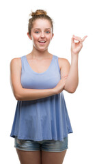 Wall Mural - Young blonde woman with a big smile on face, pointing with hand and finger to the side looking at the camera.