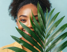 Natural Skincare Beauty Portrait Photo Of A Beautiful Young Woman Hiding Her Face Behind A Green Palm Leaf While Looking At The Camera. High-quality Adv Studio Photo Of Natural Cosmetics And Beauty