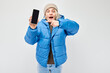 Portrait of young blond woman in blue jacket showing blank mobile phone screen with excited face. Person with smartphone isolated on white background.