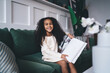 © BullRun - Joyful young girl with curly hair, wearing a cream dress, sitting on a green sofa at home, smiling while reading a large book. A cozy, educational atmosphere reflects a love for learning