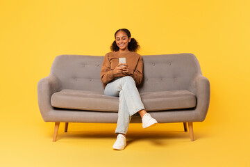 Wall Mural - Cheerful black teenager girl browsing internet on smartphone, yellow background