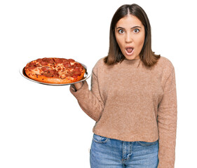 Poster - Young beautiful woman holding italian pizza scared and amazed with open mouth for surprise, disbelief face