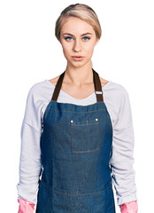 Wall Mural - Beautiful young blonde woman wearing cleaner apron and gloves with serious expression on face. simple and natural looking at the camera.