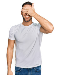 Poster - Handsome man with beard wearing casual white t shirt smiling and laughing with hand on face covering eyes for surprise. blind concept.