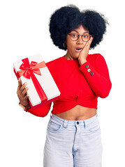 Poster - Young african american girl holding gift afraid and shocked, surprise and amazed expression with hands on face