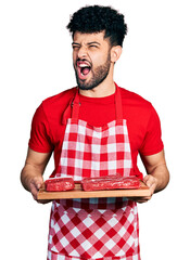 Wall Mural - Young arab man with beard holding board with raw meat angry and mad screaming frustrated and furious, shouting with anger. rage and aggressive concept.