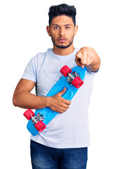 Wall Mural - Handsome latin american young man holding skate pointing with finger to the camera and to you, confident gesture looking serious