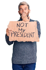 Wall Mural - Young handsome blond man holding not my president cardboard banner covering mouth with hand, shocked and afraid for mistake. surprised expression