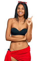 Wall Mural - Young african american woman wearing bikini with a big smile on face, pointing with hand and finger to the side looking at the camera.