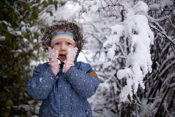 Little beautiful girl in winter clothes standing alone in the middle of a snowy forest. Girl in jacket with fur hood posing in winter forest. Shrubs and trees are covered with snow