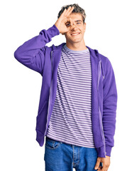 Wall Mural - Young handsome man wearing casual purple sweatshirt doing ok gesture with hand smiling, eye looking through fingers with happy face.