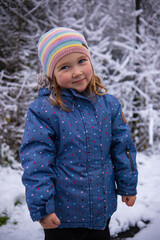 Wall Mural - Little beautiful girl in winter clothes standing alone in the middle of a snowy forest. Girl in jacket with fur hood posing in winter forest. Shrubs and trees are covered with snow