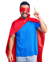 Canvas Print - Young hispanic man wearing super hero costume pointing finger up with successful idea. exited and happy. number one.