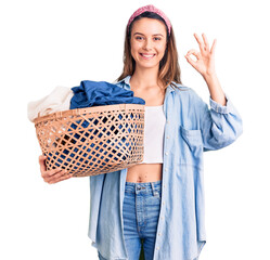 Canvas Print - Young beautiful girl holding laundry basket doing ok sign with fingers, smiling friendly gesturing excellent symbol