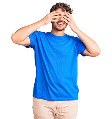 Wall Mural - Young handsome man with curly hair wearing casual clothes covering eyes with hands smiling cheerful and funny. blind concept.