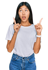 Poster - Beautiful young asian woman wearing casual white t shirt amazed and surprised looking up and pointing with fingers and raised arms.