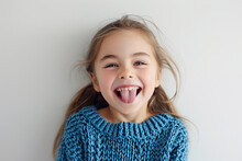 A cute Caucasian girl in a blue knitted sweater against a white wall with a happy and funny face, smiling and sticking out her tongue