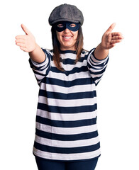 Poster - Young beautiful brunette woman wearing burglar mask looking at the camera smiling with open arms for hug. cheerful expression embracing happiness.