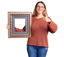 Poster - Beautiful young woman holding empty frame smiling happy and positive, thumb up doing excellent and approval sign