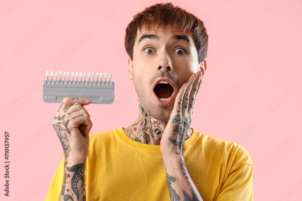 Shocked young tattooed man with teeth color samples on pink background, closeup