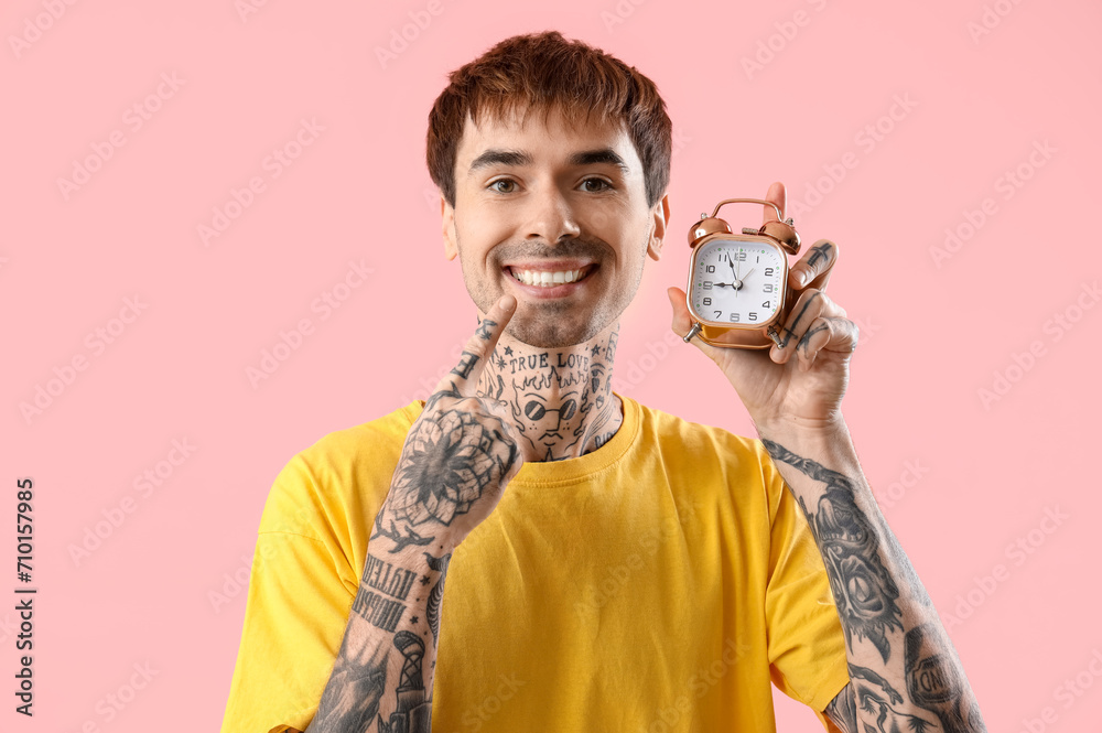 Young tattooed man with alarm clock pointing at his teeth on pink background