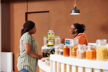 Wall Mural - Vendor assisting female client with nutritious products. Cashier at eco friendly store promotes healthy lifestyle, offering fresh locally grown organic produce and sustainable, zero waste options.