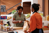 Fototapeta Na drzwi - Female consumer paying with credit card at checkout counter, purchasing organic pantry staples at local supermarket. African american women at cashier desk using pos machine for seamless transactions.