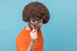© khosrork - Funny man with Afro hairstyle wearing orange T-shirt standing, holding magnifying glass and looking at camera with big zoom eye and funny expression. Indoor studio shot isolated on blue background.