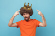 © khosrork - Portrait of self confident man with Afro hairstyle wearing orange T-shirt standing and pointing at golden crown, looking at camera with proud expression. Indoor studio shot isolated on blue background