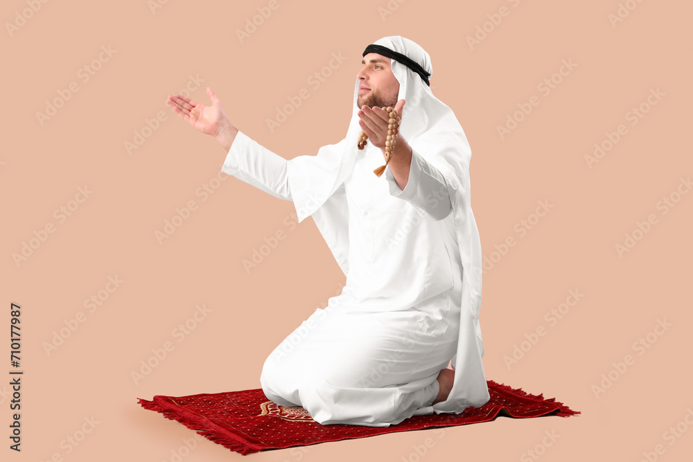 Young Muslim man praying on mat against beige background. Ramadan celebration