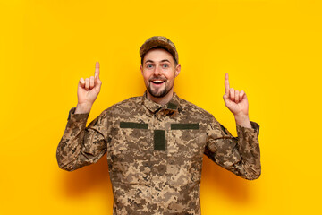 young Ukrainian male soldier in camouflage pixel uniform points his hands up on a yellow isolated background, guy military cadet of the Ukrainian army advertises copy space from above