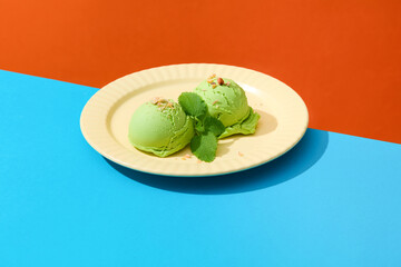 Two matcha ice cream balls with basil leaves and crushed nuts are displayed on a round ceramic plate. The background is divided into two colors orange and blue.