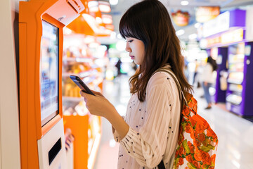 Young Asian woman using a touchscreen self-service kiosk while holding a smartphone in a shopping mall.