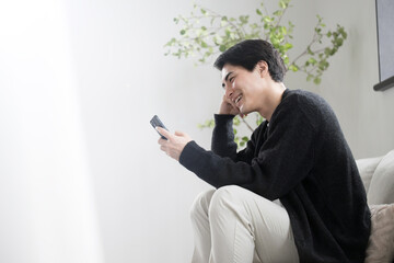 綺麗な白いリビングでスマホを見るくつろぐ長袖の男性の横顔 スマホを見て考える NISAや投資ってどうする？イメージ