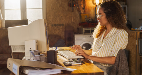 Fototapete - Side View Of Hispanic Female Engineering Student Writing Academic Thesis On Old Desktop Computer In Retro Garage. Future Engineer Focused On Innovative Project For University Of Technology In Nineties
