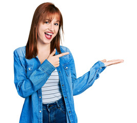 Canvas Print - Redhead young woman wearing casual denim jacket amazed and smiling to the camera while presenting with hand and pointing with finger.