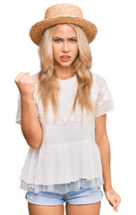 Poster - Young blonde girl wearing summer hat angry and mad raising fist frustrated and furious while shouting with anger. rage and aggressive concept.