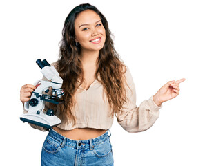 Wall Mural - Young hispanic girl holding microscope smiling happy pointing with hand and finger to the side