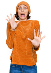 Poster - Young hispanic woman wearing casual clothes and wool cap afraid and terrified with fear expression stop gesture with hands, shouting in shock. panic concept.