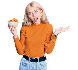Wall Mural - Young caucasian woman holding nachos potato chips celebrating achievement with happy smile and winner expression with raised hand