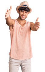 Wall Mural - Young hispanic man wearing summer hat looking at the camera smiling with open arms for hug. cheerful expression embracing happiness.