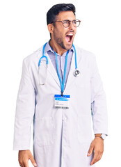 Wall Mural - Young hispanic man wearing doctor uniform and stethoscope angry and mad screaming frustrated and furious, shouting with anger. rage and aggressive concept.