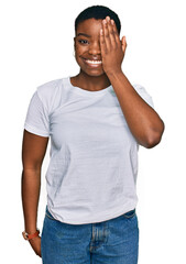 Canvas Print - Young african american woman wearing casual white t shirt covering one eye with hand, confident smile on face and surprise emotion.